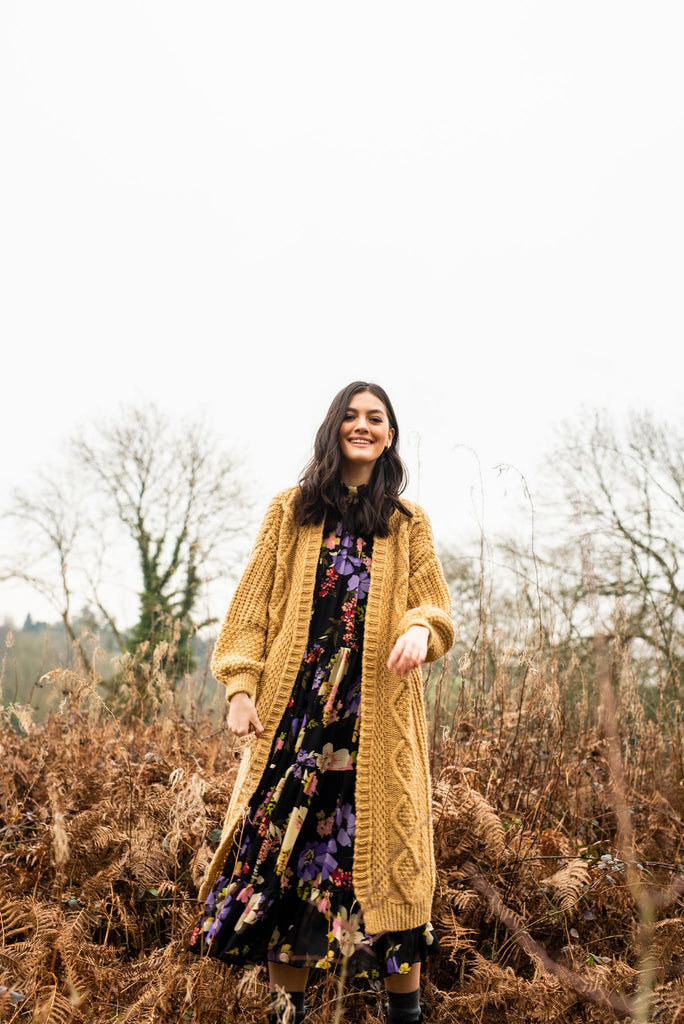 Smiling woman stood outside on a winters day wearing a long mustard yellow cardigan and long dress 