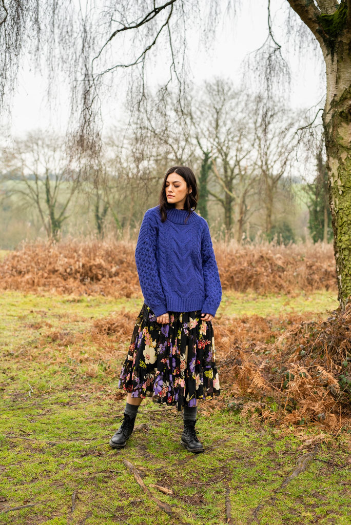 Full length photo of a woman standing outside beside a tree wearing a dress and violet blue cable knit jumper