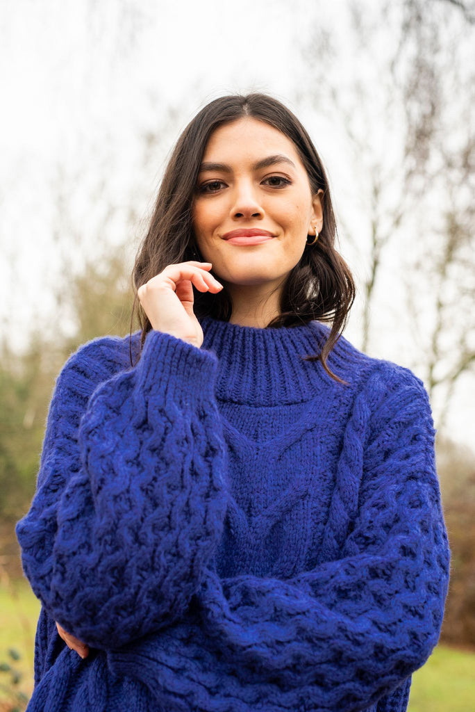 Portrait photo of a woman standing outside and smiling, wearing a violet blue chunky knit jumper