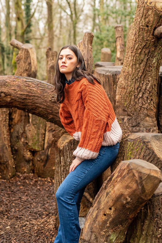 Woman in an orange jumper and blue jeans leaning against a tree