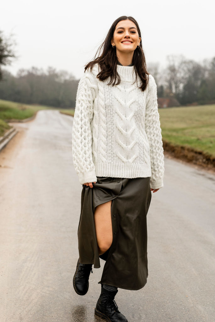 Smiling woman walking on a rural road toward the camera, wearing a long dark skirt a white cable jumper