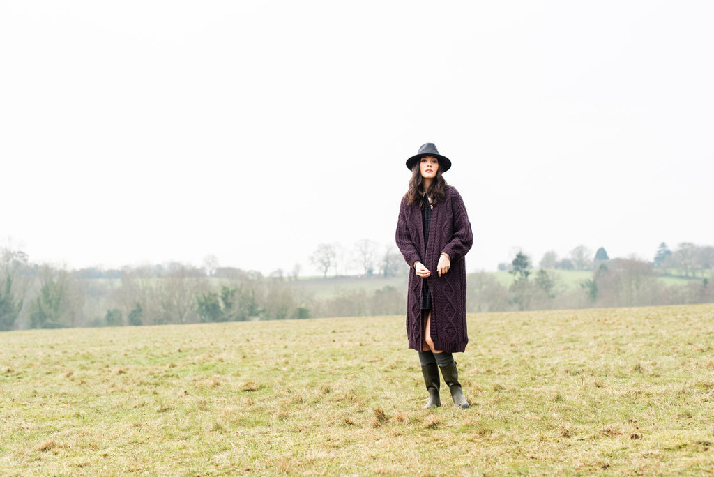 Woman standing mid-ground on grass with fields and trees in the background, wearing a dark hat and long brown cardigan