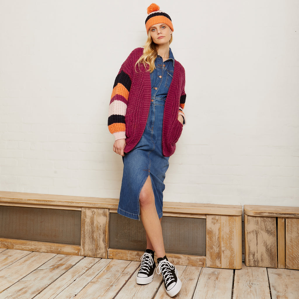 Full length photo of a woman in a denim dress, yellow bobble hat and the Allie berry cardigan leaning against a wall