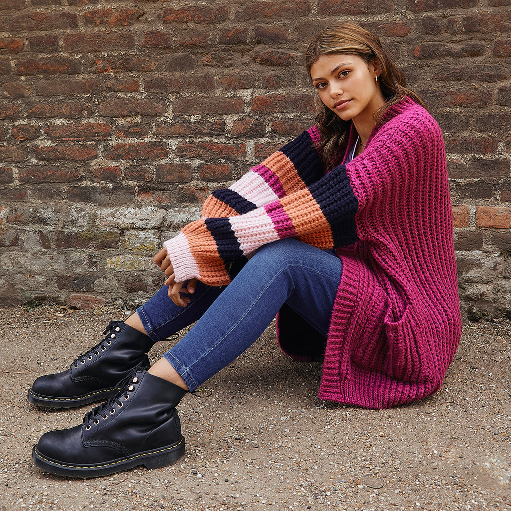 Woman sat on the ground in front of a stone wall, leaning on her legs, wearing black boots, blue jeans and the berry stripe Allie cardigan