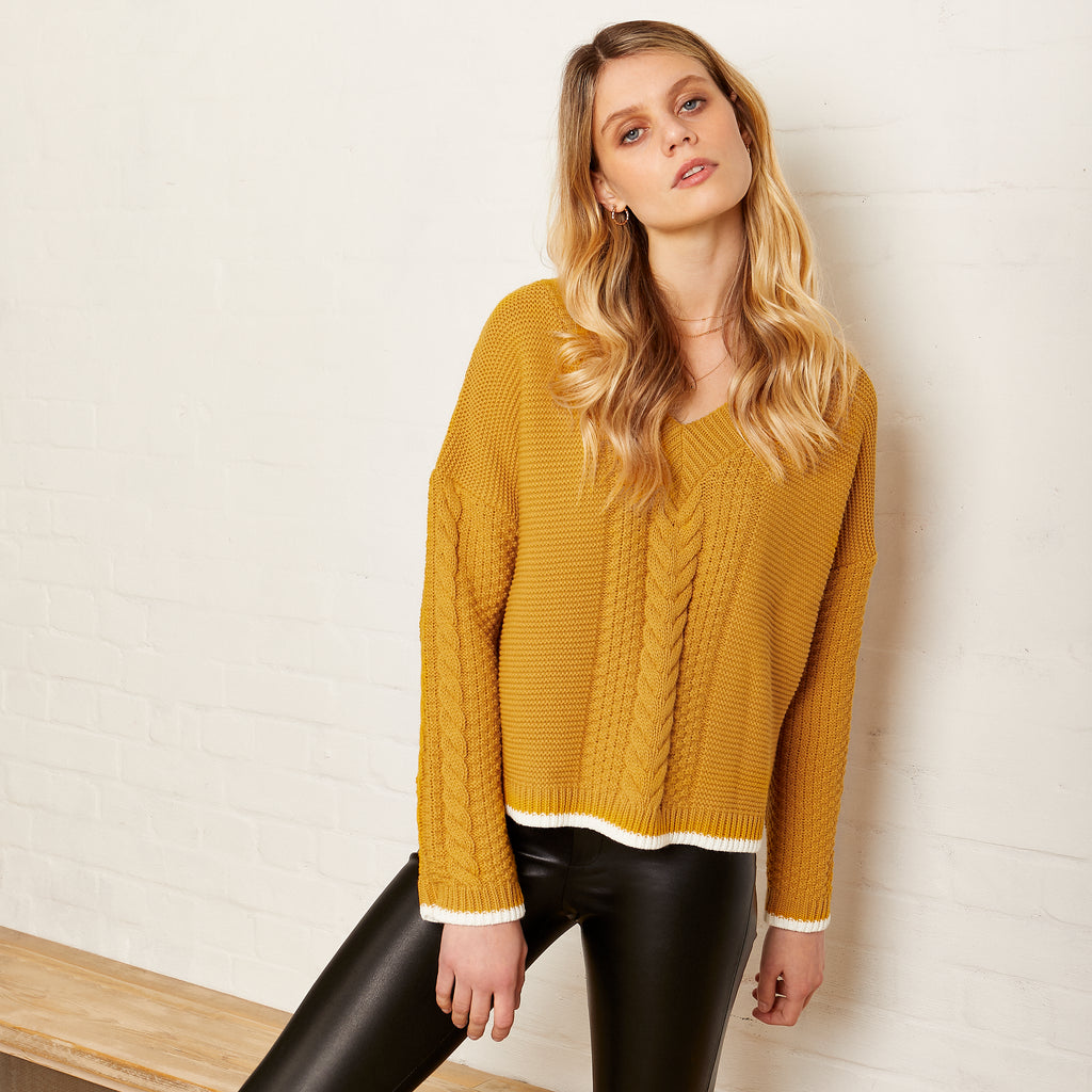 Woman leaning against a white brick wall wearing Emma mustard yellow knitted jumper