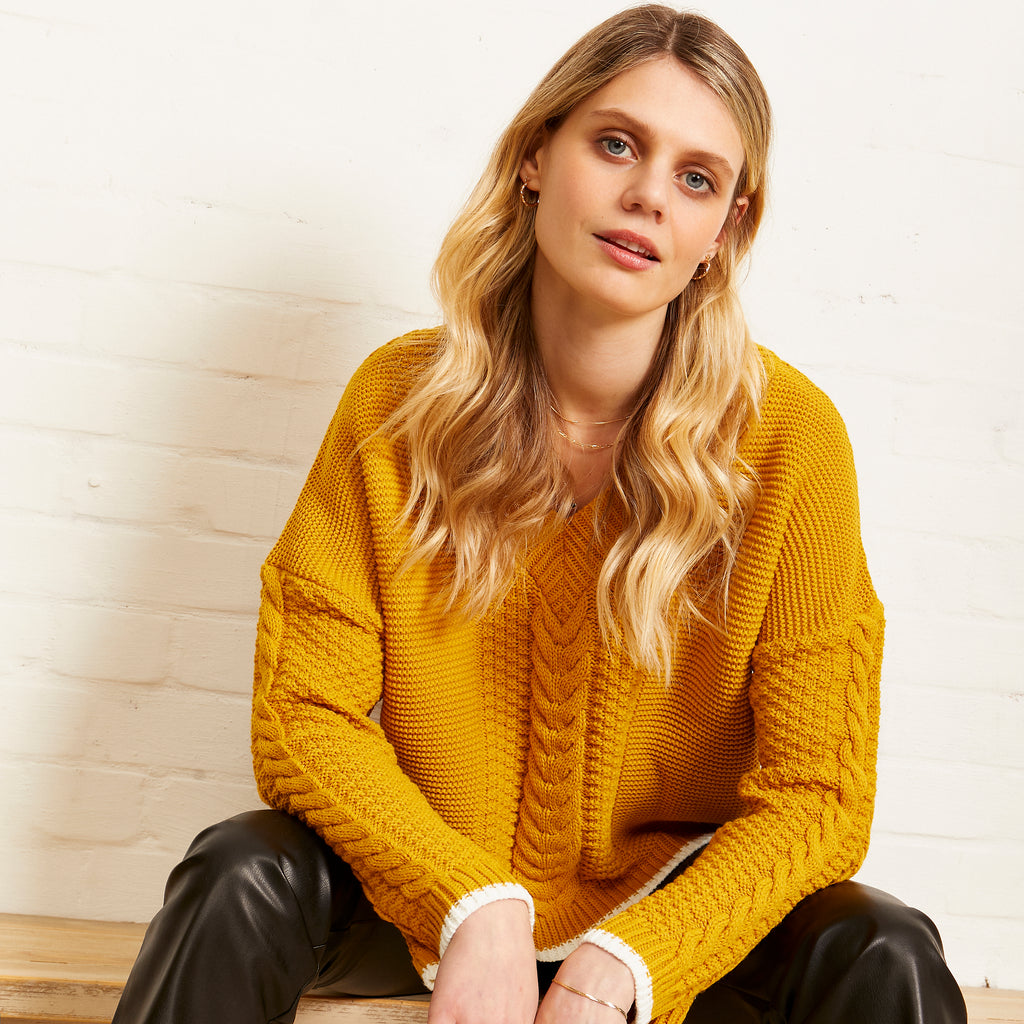 Woman sat and leaning face on, wearing the Emma mustard yellow knitted jumper