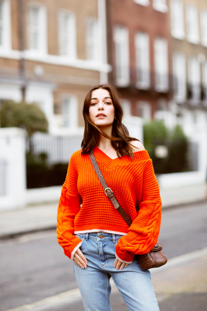 A woman standing in a suburban street, wearing a burnt orange knitted jumper with brown shoulder bag