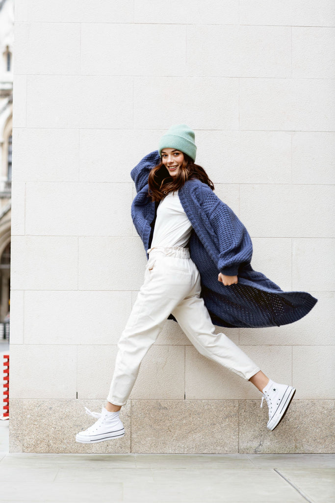 Action shot of woman running wearing a long navy cardigan, white trousers and a knitted mint hat