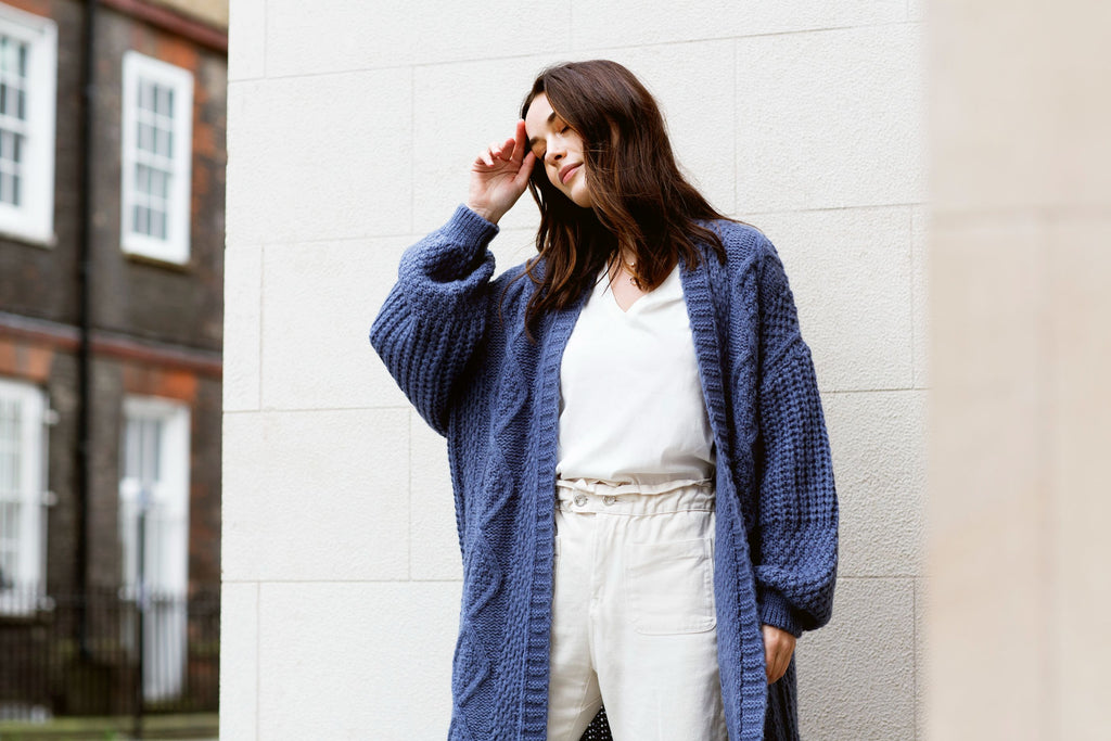 Woman with closed eyes standing outside a white building wearing a navy blue long knitted cardigan over a white outfit