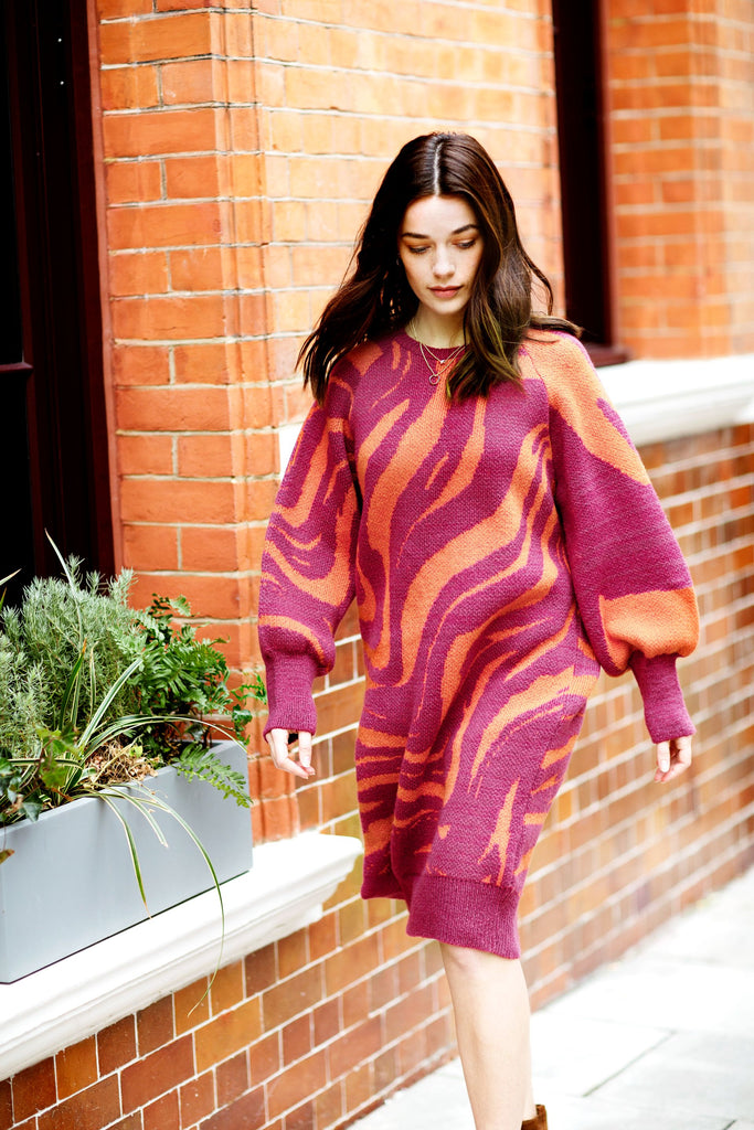 Woman walking past a brick building with head down, wearing a raspberry red and orange knitted jumper dress