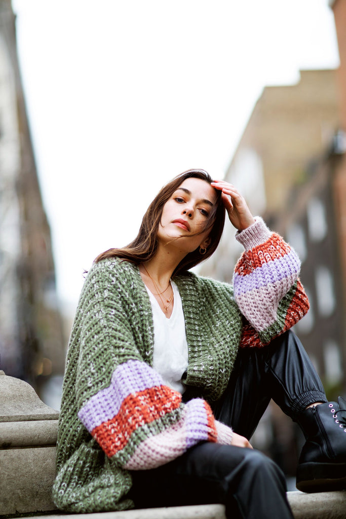 Woman sitting outside on a stone bench wearing a khaki green cardigan with lilac and orange stripes