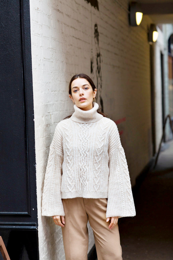 Lady standing beside a wall wearing a white roll neck cable jumper