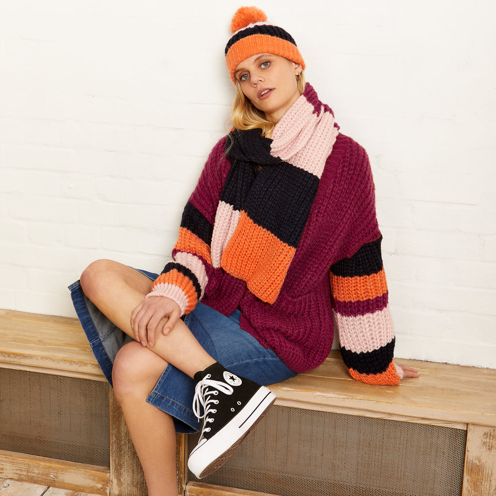 Woman sat on a wooden bench wearing a berry purple orange stripe knitted cardigan with matching hat and scarf